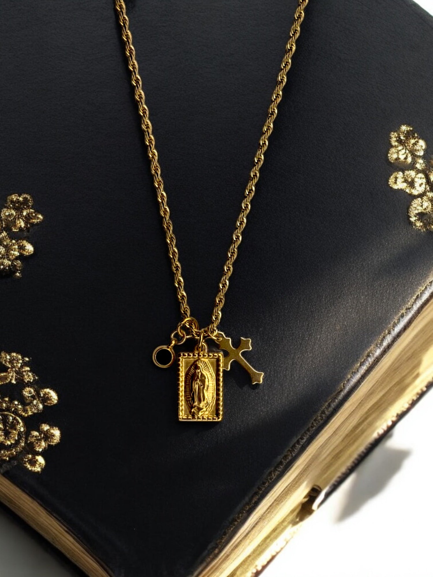 Elegant gold religious necklace with layered charms, including a Virgin Mary pendant, a baroque-style cross, and a small black gemstone accent.